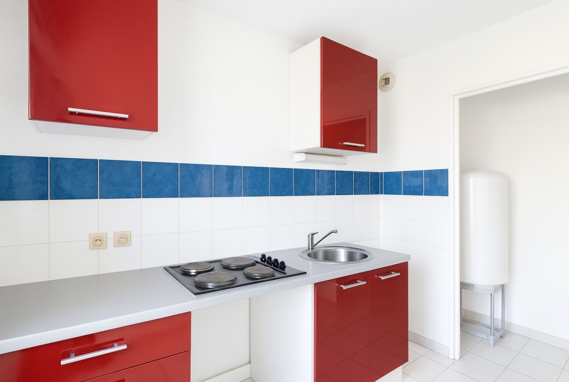 Cuisine avec plan de travail clair, plaques électriques et évier inox. Des rangements rouges et des carreaux blancs et bleus composent l’espace.
