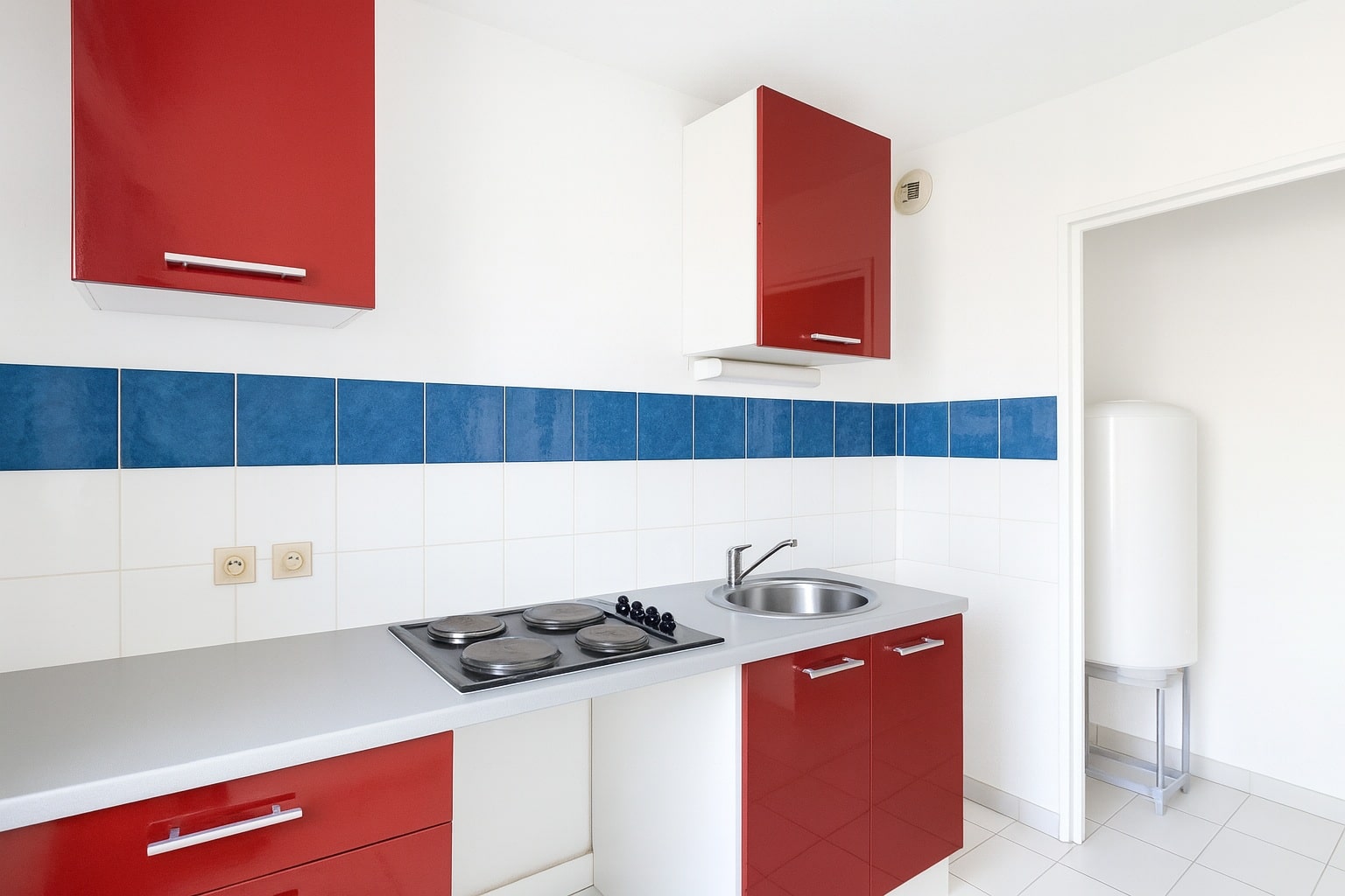 Cuisine avec plan de travail clair, plaques électriques et évier inox. Des rangements rouges et des carreaux blancs et bleus composent l’espace.