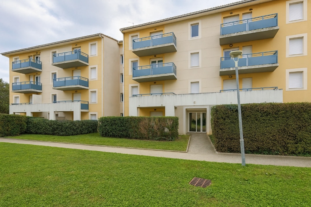 Façade beige et jaune d’une résidence avec plusieurs balcons bleus. Des espaces verts et des haies aménagées entourent les bâtiments.