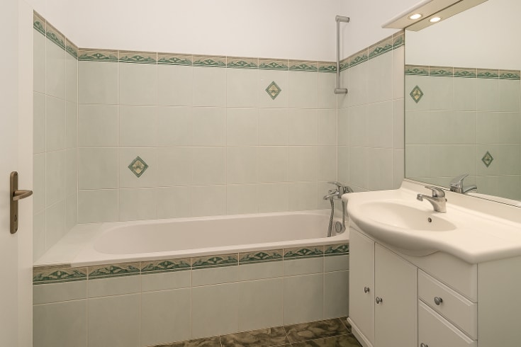 Salle de bains équipée d’une baignoire encastrée et d’un meuble vasque blanc. L’espace est entièrement carrelé avec des motifs verts et dispose d’un éclairage intégré au-dessus du miroir.