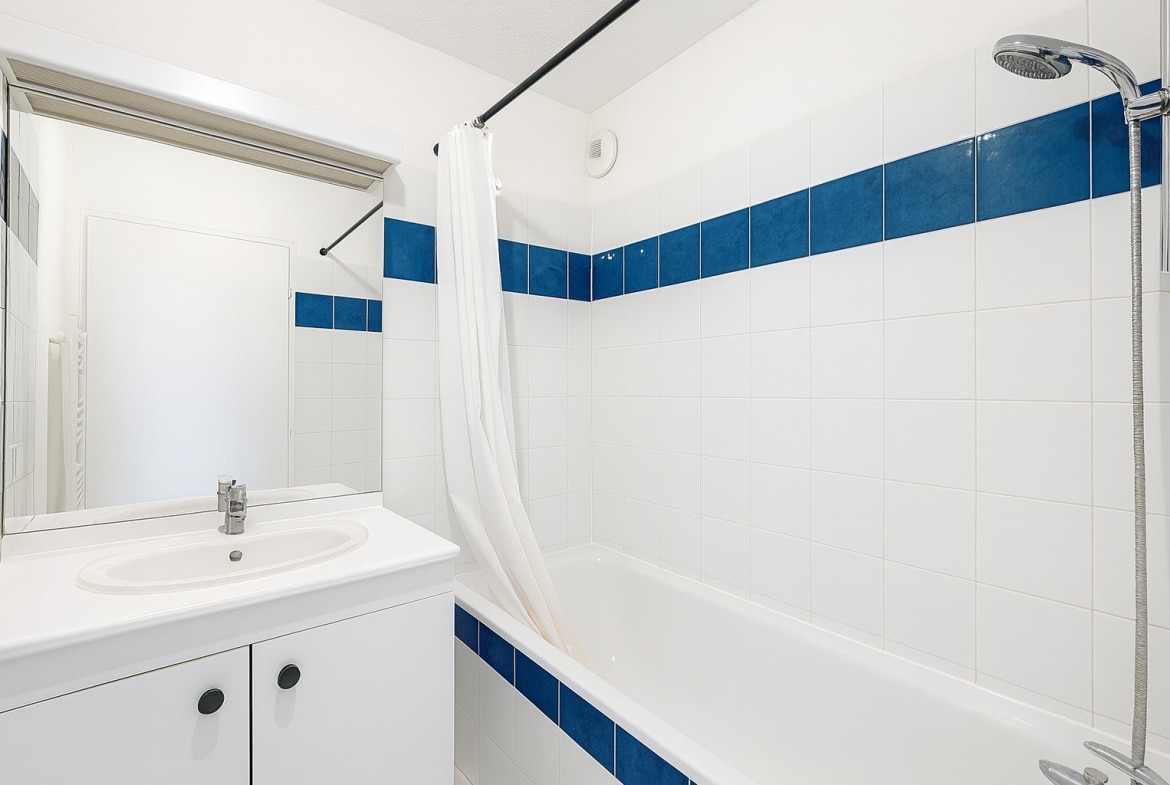 Salle de bain comprenant une baignoire entourée de faïence blanche et bleue. Un meuble vasque blanc et un miroir complètent la pièce.