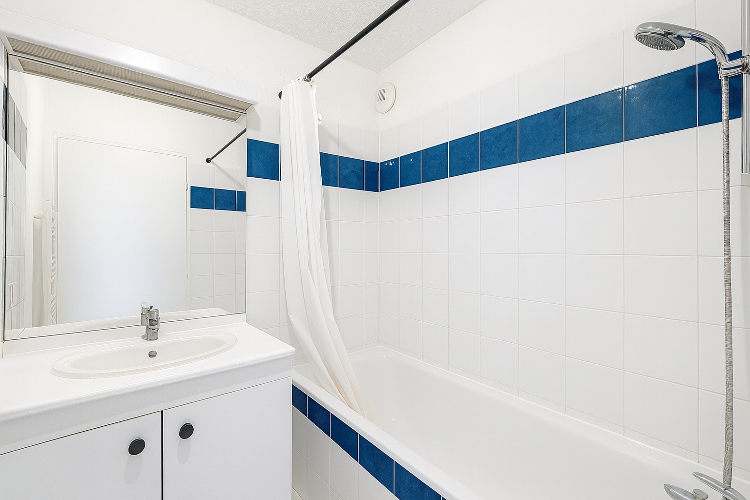 Salle de bain comprenant une baignoire entourée de faïence blanche et bleue. Un meuble vasque blanc et un miroir complètent la pièce.