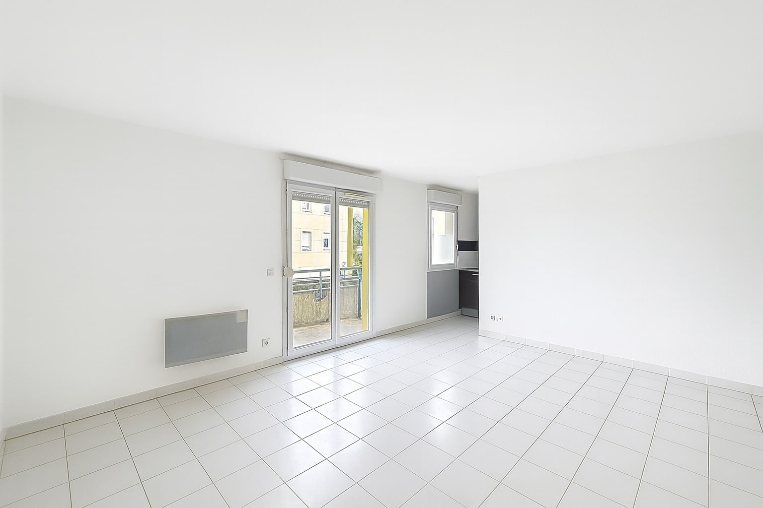 Salon entièrement carrelé et peint en blanc avec une grande baie vitrée. L’espace s’ouvre sur une terrasse extérieure offrant une vue dégagée.