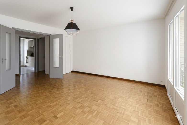 Séjour spacieux avec parquet en mosaïque et doubles portes vitrées. La pièce bénéficie d’une grande luminosité grâce aux ouvertures donnant sur l’extérieur.