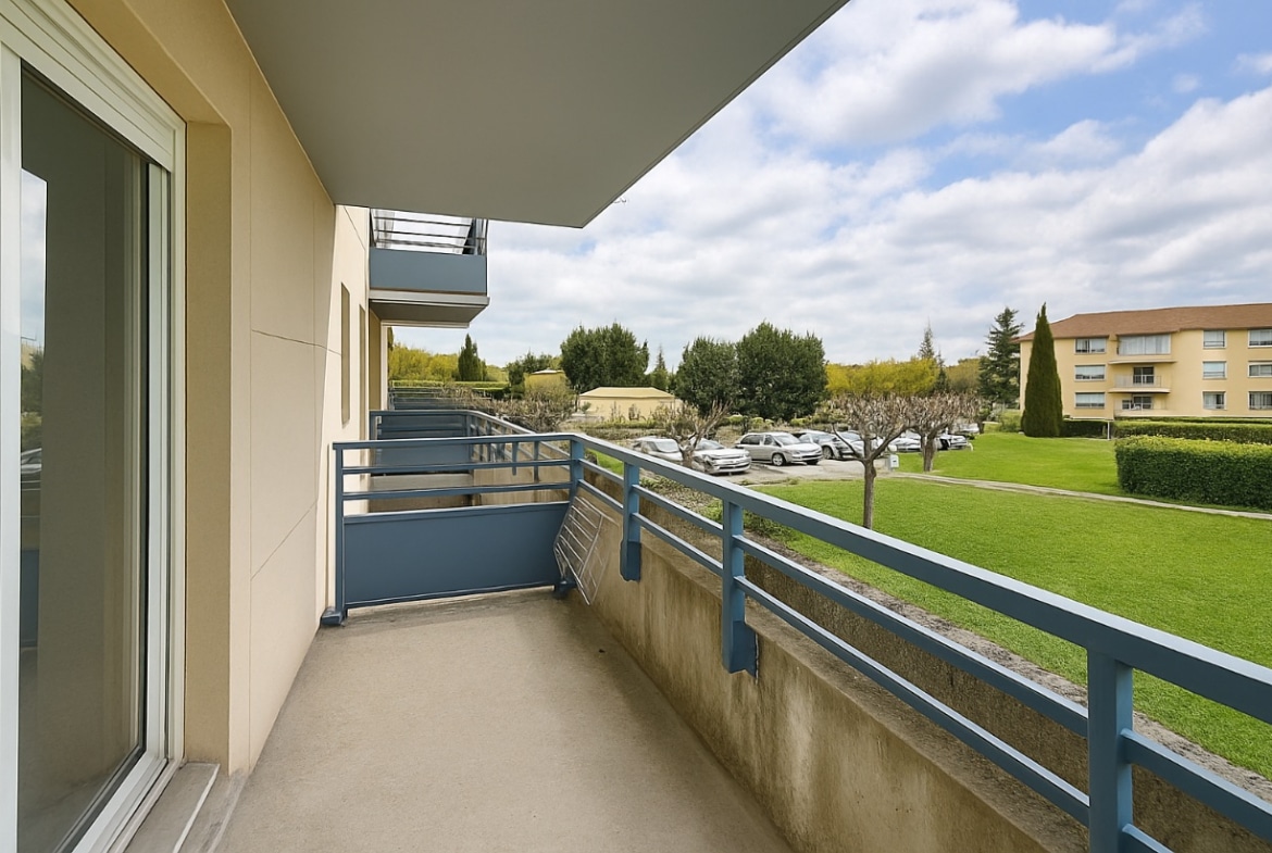 Terrasse avec garde-corps bleus offrant une vue sur les jardins et le parking résidentiel. L’espace est accessible depuis la pièce de vie par une baie vitrée
