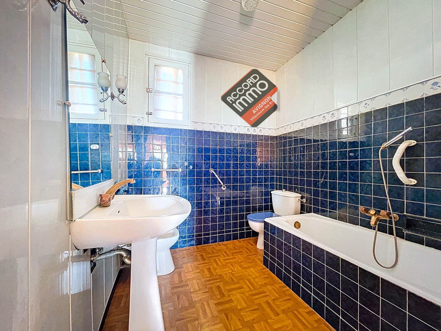 Salle de bain lumineuse et moderne avec carrelage bleu et sol en parquet, équipée d'une baignoire, d'un lavabo et de toilettes située dans une maison avec jardin à vendre à Avignon