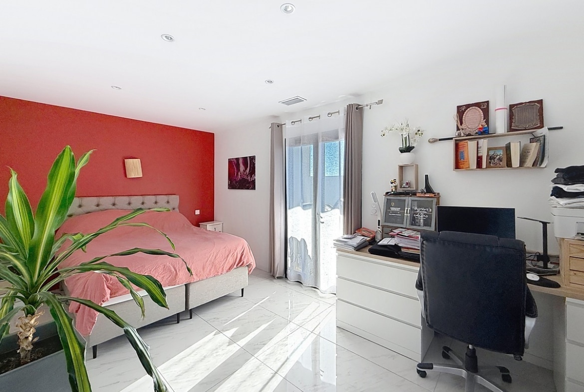 Chambre avec lit double à tête de lit capitonnée grise et linge de lit rose, mur d'accent peint en rouge vif. Espace bureau avec chaise de travail noire, commode blanche et étagères murales, plante verte en premier plan et porte-fenêtre avec voilage.