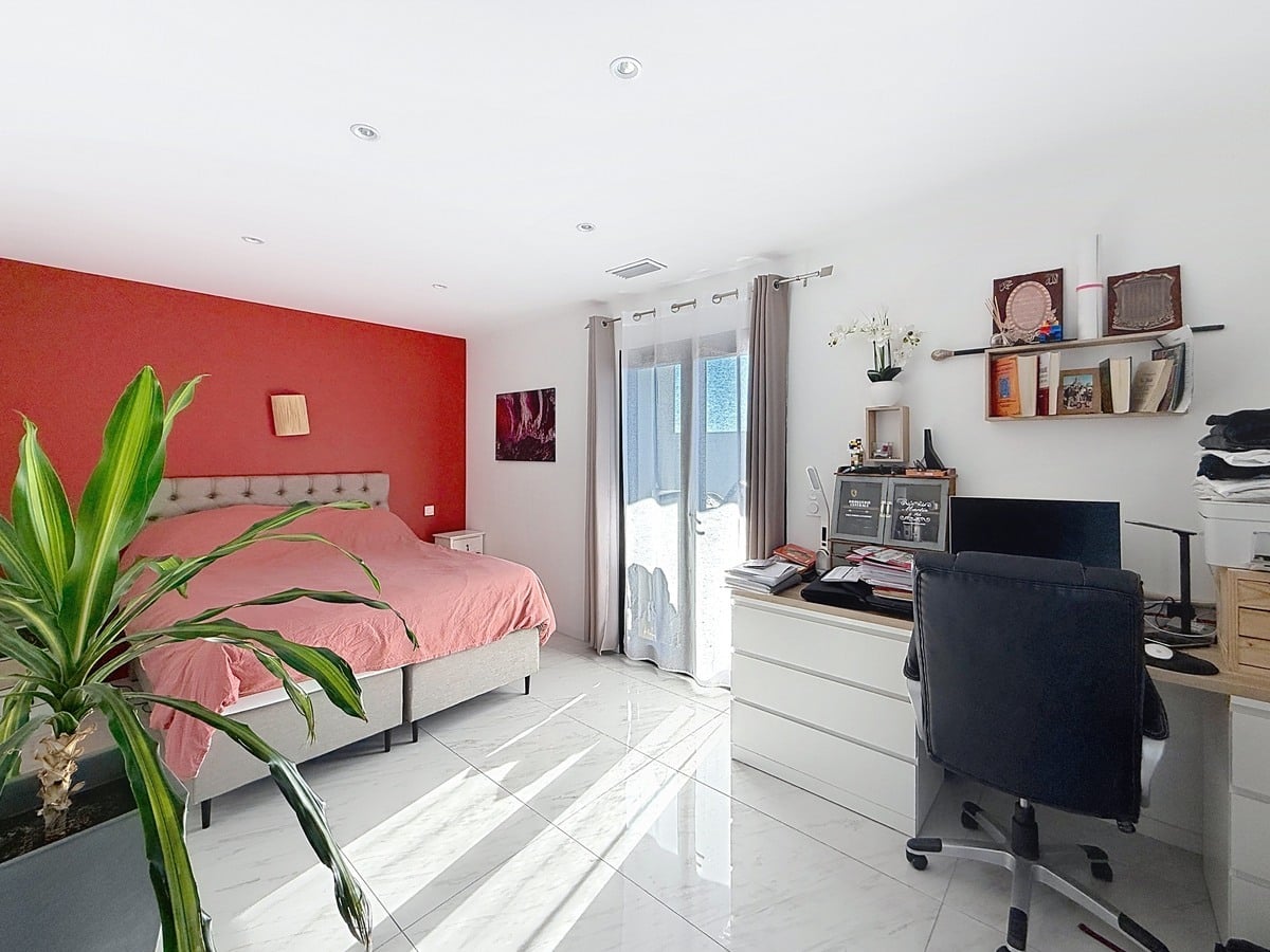 Chambre avec lit double à tête de lit capitonnée grise et linge de lit rose, mur d'accent peint en rouge vif. Espace bureau avec chaise de travail noire, commode blanche et étagères murales, plante verte en premier plan et porte-fenêtre avec voilage.