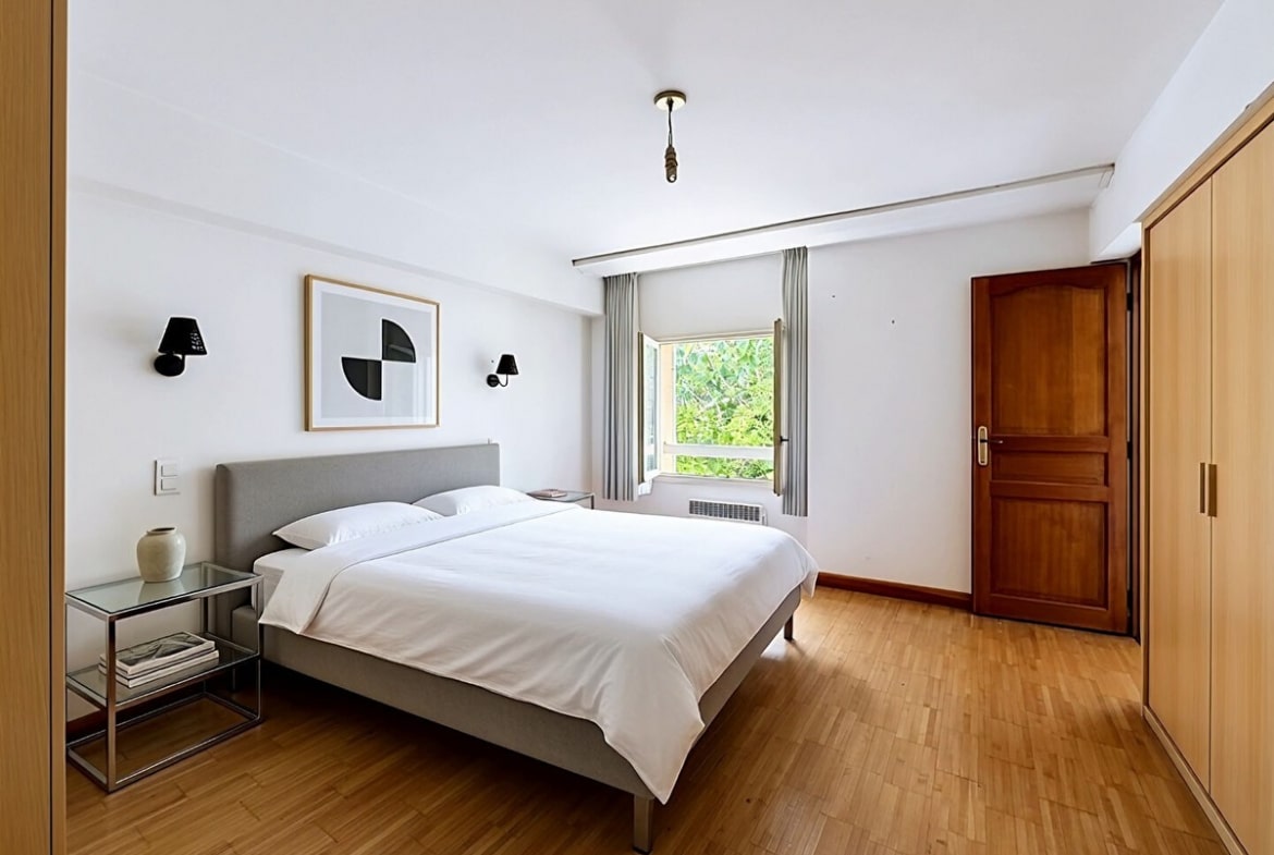 Chambre avec lit double à tête de lit grise et linge blanc, parquet en bois clair au sol. Placard intégré avec portes en bois clair et porte d'entrée en bois foncé, fenêtre avec rideaux gris donnant sur la végétation.