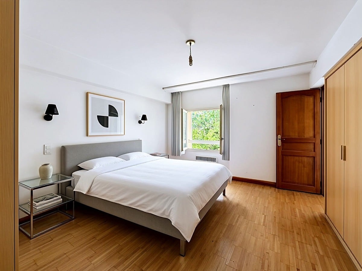Chambre avec lit double à tête de lit grise et linge blanc, parquet en bois clair au sol. Placard intégré avec portes en bois clair et porte d'entrée en bois foncé, fenêtre avec rideaux gris donnant sur la végétation.