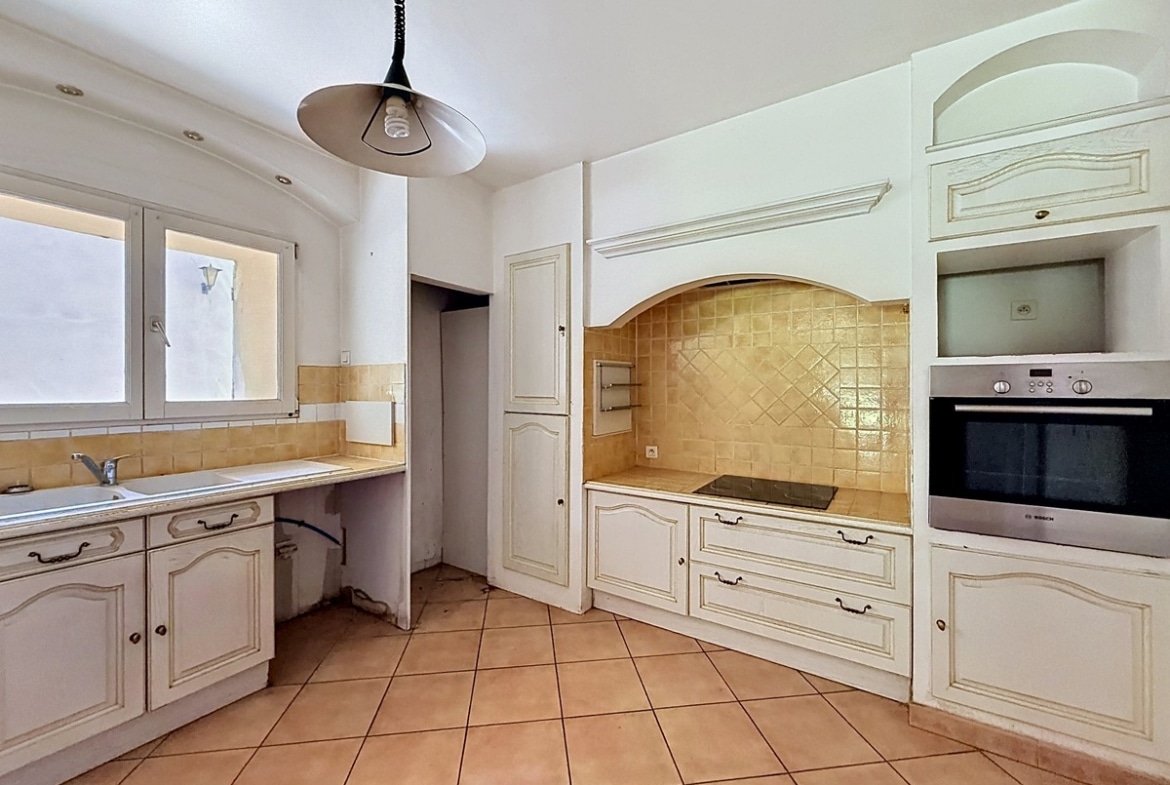 Cuisine avec meubles blancs à portes moulurées et crédence en carrelage jaune ocre. Four encastré inox, plaque de cuisson vitrocéramique, évier sous fenêtre et sol en carrelage terre cuite.