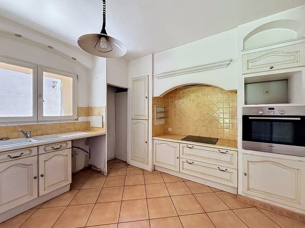 Cuisine avec meubles blancs à portes moulurées et crédence en carrelage jaune ocre. Four encastré inox, plaque de cuisson vitrocéramique, évier sous fenêtre et sol en carrelage terre cuite.