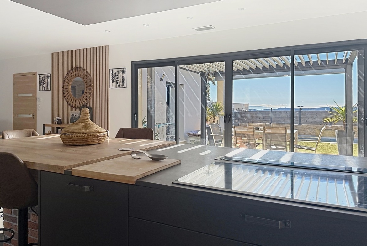 Plan de travail de cuisine gris avec plaque à induction et planche à découper en bois. Panneau mural en tasseaux de bois avec miroir soleil en rotin, baies vitrées coulissantes révélant une terrasse avec table et chaises sous pergola.
