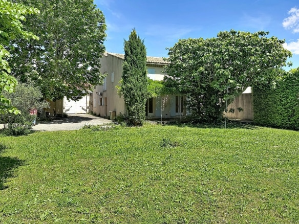 Maison provençale à deux niveaux avec enduit beige et volets clairs, entourée de végétation dense incluant cyprès, platanes et haie de lierre. Pelouse au premier plan légèrement haute avec jeune arbre planté au centre du terrain.
