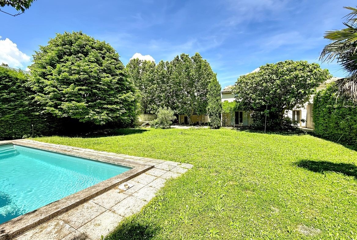Grand jardin avec pelouse verte et nombreux arbres matures formant un écrin de verdure. Piscine rectangulaire avec margelles en pierre et vue sur la façade de la maison en arrière-plan.
