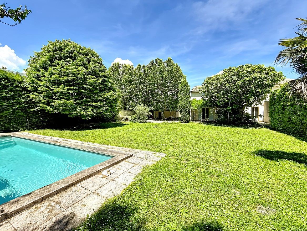 Grand jardin avec pelouse verte et nombreux arbres matures formant un écrin de verdure. Piscine rectangulaire avec margelles en pierre et vue sur la façade de la maison en arrière-plan.