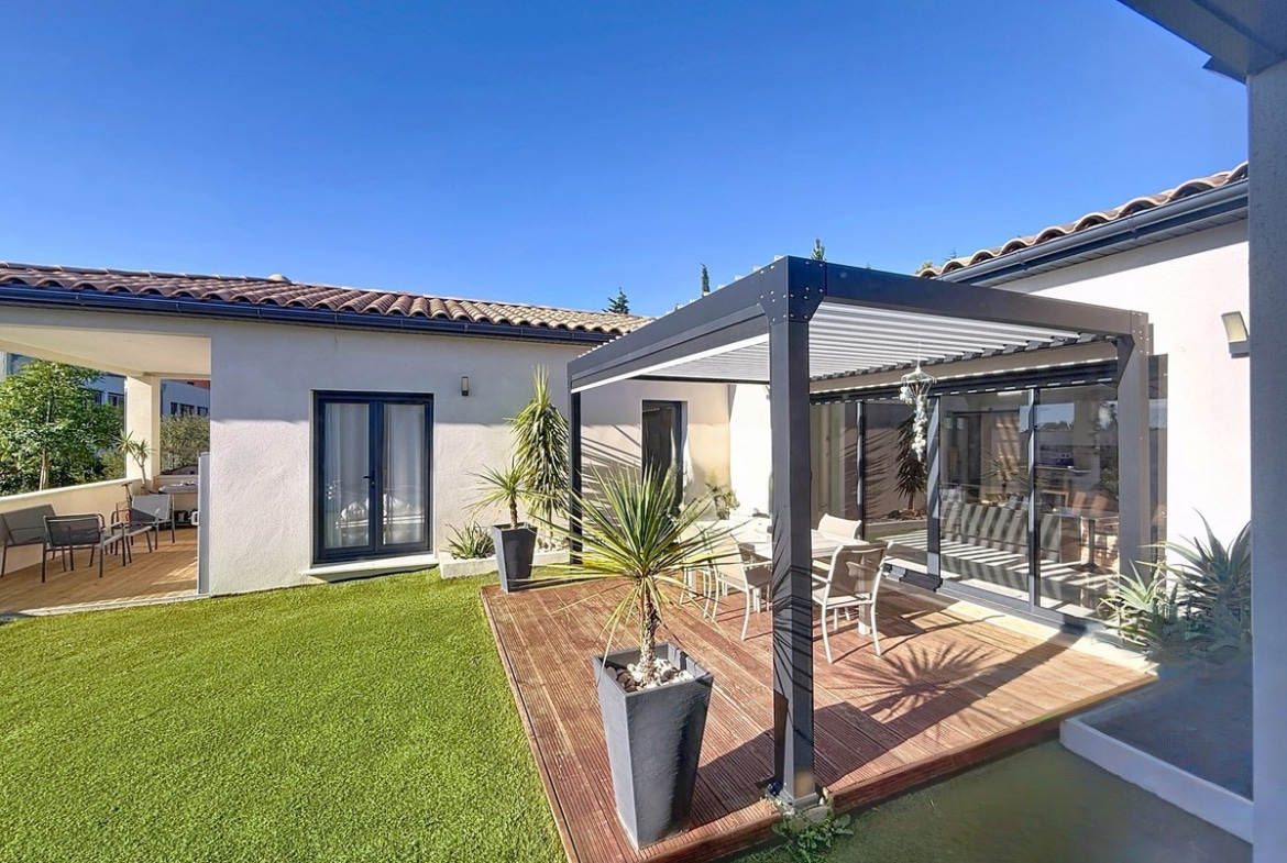 Jardin avec pelouse verte et terrasse en bois sous pergola bioclimatique à lames orientables grises. Maison de plain-pied avec façade blanche, tuiles canal traditionnelles, menuiseries anthracite et palmiers en pots décoratifs.