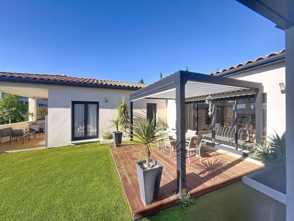 Jardin avec pelouse verte et terrasse en bois sous pergola bioclimatique à lames orientables grises. Maison de plain-pied avec façade blanche, tuiles canal traditionnelles, menuiseries anthracite et palmiers en pots décoratifs.