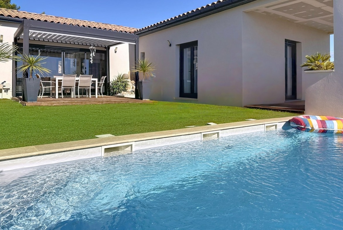 Piscine rectangulaire à eau bleue avec bouée multicolore flottant à la surface, bordée de margelles claires. Jardin avec pelouse verte, terrasse en bois sous pergola avec salon de jardin blanc et maison de plain-pied aux murs blancs et gris.