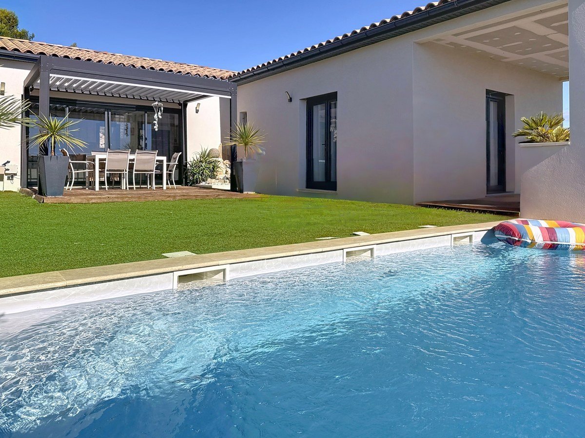 Piscine rectangulaire à eau bleue avec bouée multicolore flottant à la surface, bordée de margelles claires. Jardin avec pelouse verte, terrasse en bois sous pergola avec salon de jardin blanc et maison de plain-pied aux murs blancs et gris.
