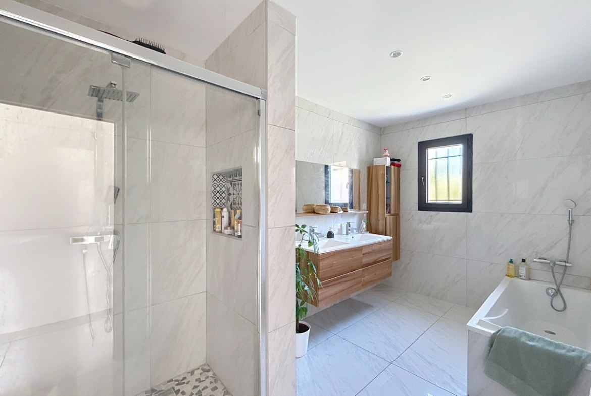 Salle de bains avec carrelage mural effet marbre blanc, douche italienne à paroi vitrée coulissante et sol en galets. Meuble vasque suspendu en bois clair, baignoire rectangulaire blanche et fenêtre à cadre noir.