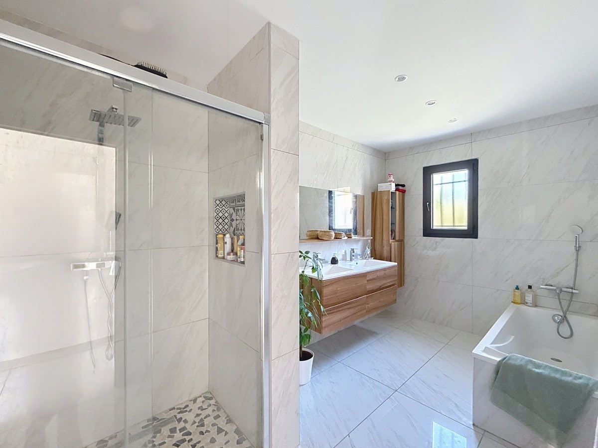 Salle de bains avec carrelage mural effet marbre blanc, douche italienne à paroi vitrée coulissante et sol en galets. Meuble vasque suspendu en bois clair, baignoire rectangulaire blanche et fenêtre à cadre noir.