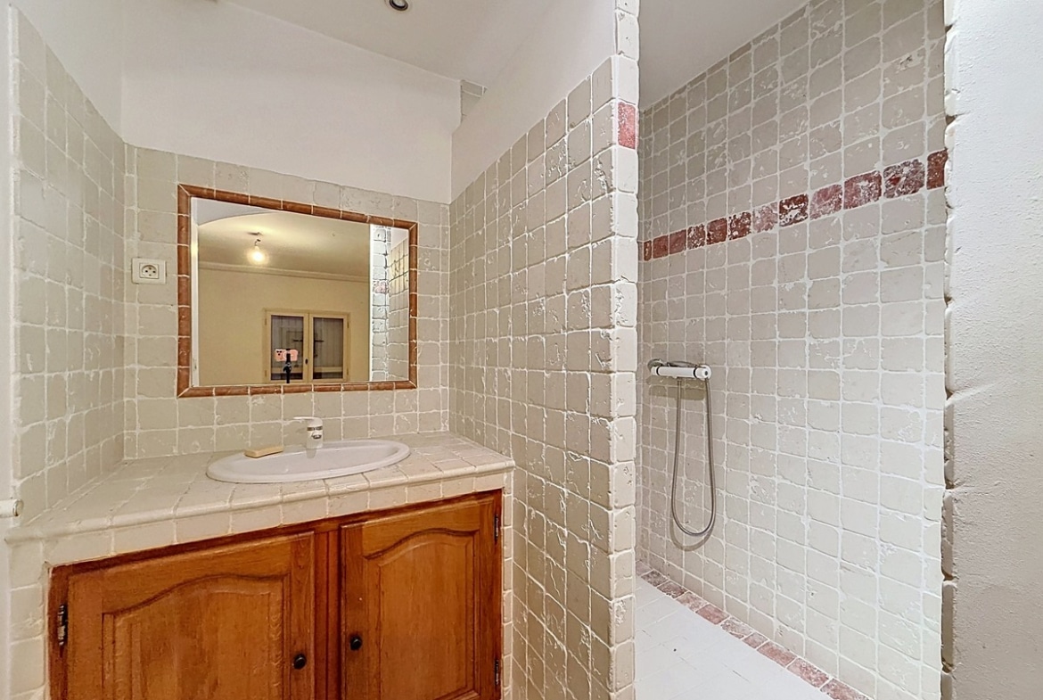 Salle d'eau avec murs en carrelage beige et frise décorative bordeaux. Meuble vasque en bois avec plan en carrelage, miroir encadré et douche à l'italienne avec robinetterie chromée.