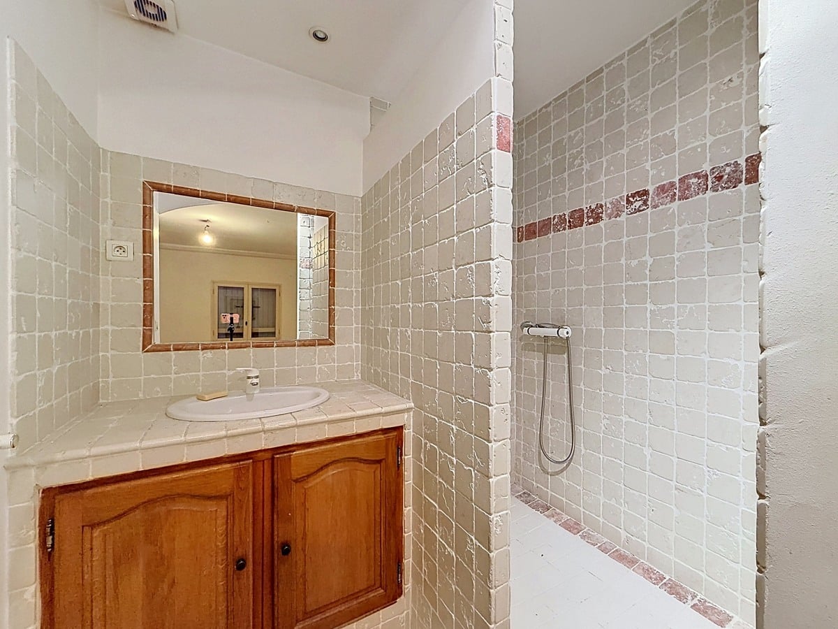 Salle d'eau avec murs en carrelage beige et frise décorative bordeaux. Meuble vasque en bois avec plan en carrelage, miroir encadré et douche à l'italienne avec robinetterie chromée.