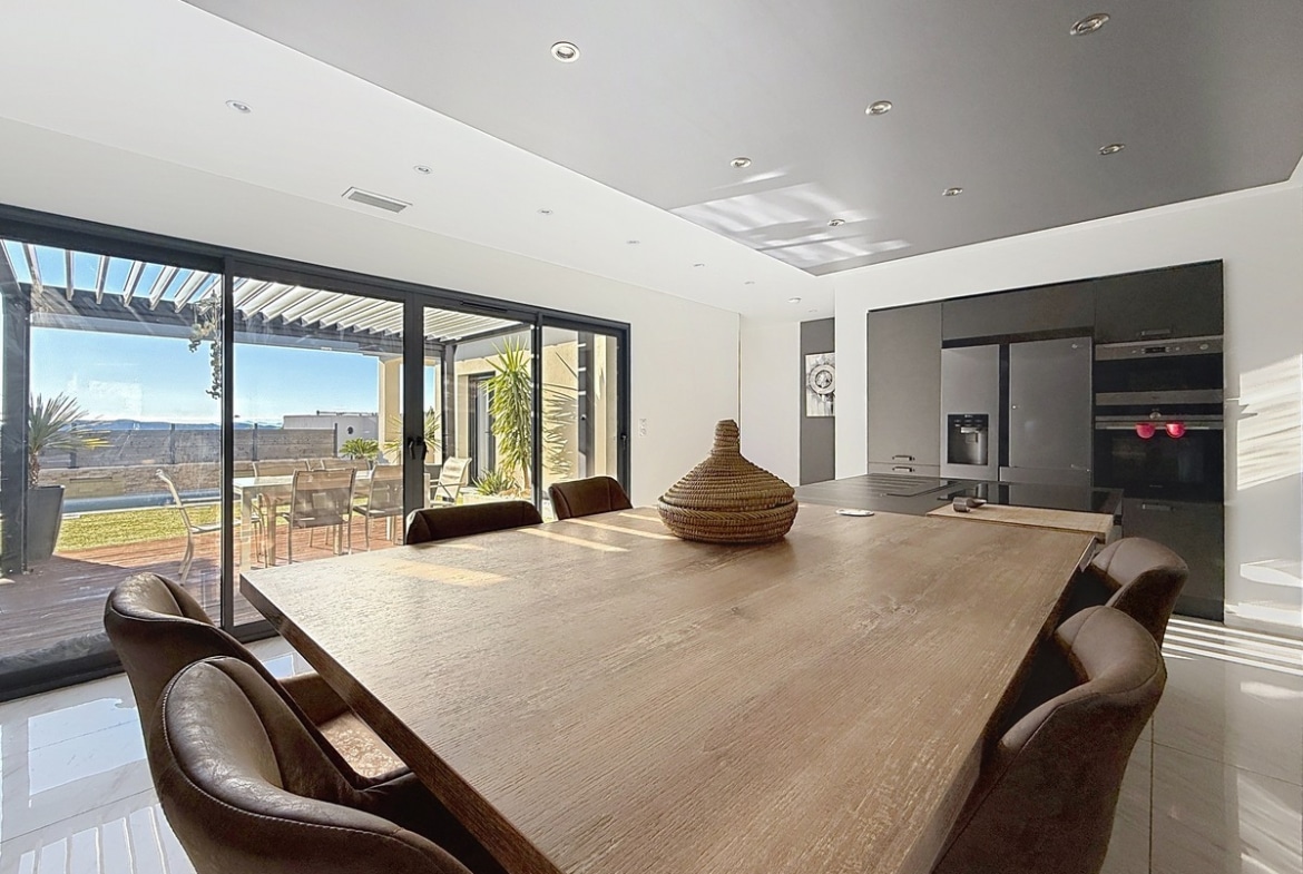 Table à manger en bois massif avec fauteuils en cuir marron et panier décoratif tressé. Grandes baies vitrées coulissantes à cadre noir ouvrant sur une terrasse couverte avec mobilier de jardin et vue sur paysage vallonné.