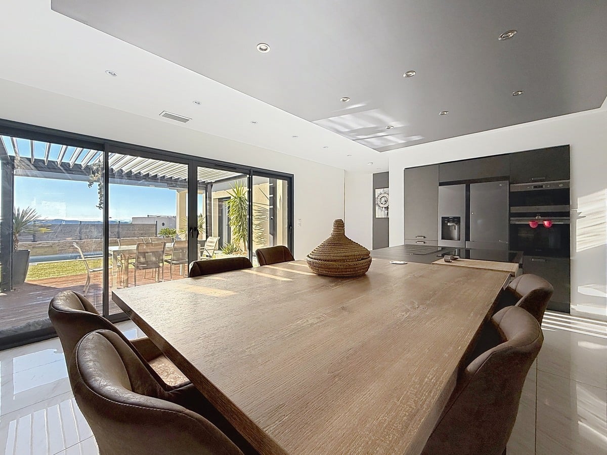 Table à manger en bois massif avec fauteuils en cuir marron et panier décoratif tressé. Grandes baies vitrées coulissantes à cadre noir ouvrant sur une terrasse couverte avec mobilier de jardin et vue sur paysage vallonné.