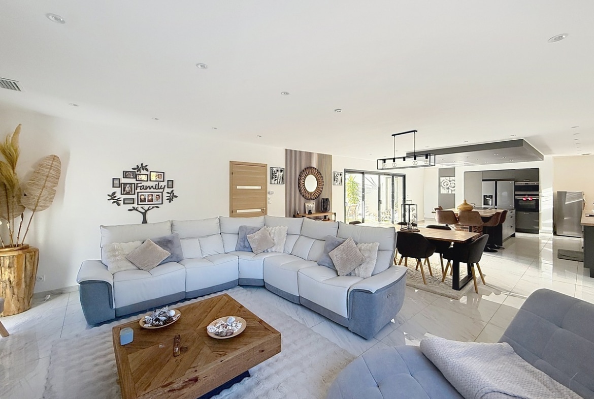 Vaste séjour ouvert avec canapé d'angle gris clair, table basse en bois à motif chevron et sol en carrelage blanc. Décoration murale en forme d'arbre généalogique avec cadres photos, panneau en tasseaux de bois verticaux et miroir rond en rotin visible en arrière-plan.