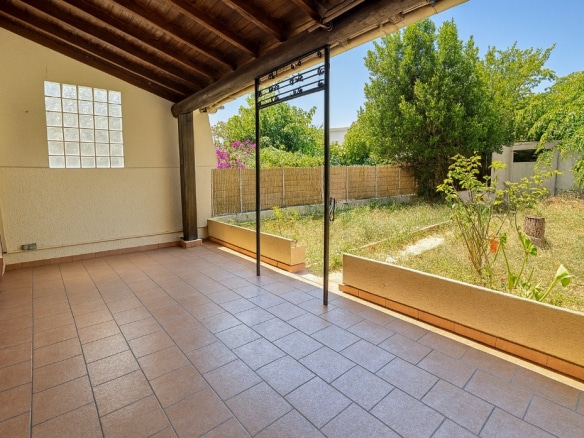 Terrasse couverte avec sol carrelé et structure en bois ouvrant sur un jardin privatif. Le jardin dispose d’arbustes et d’une clôture végétale assurant l’intimité.