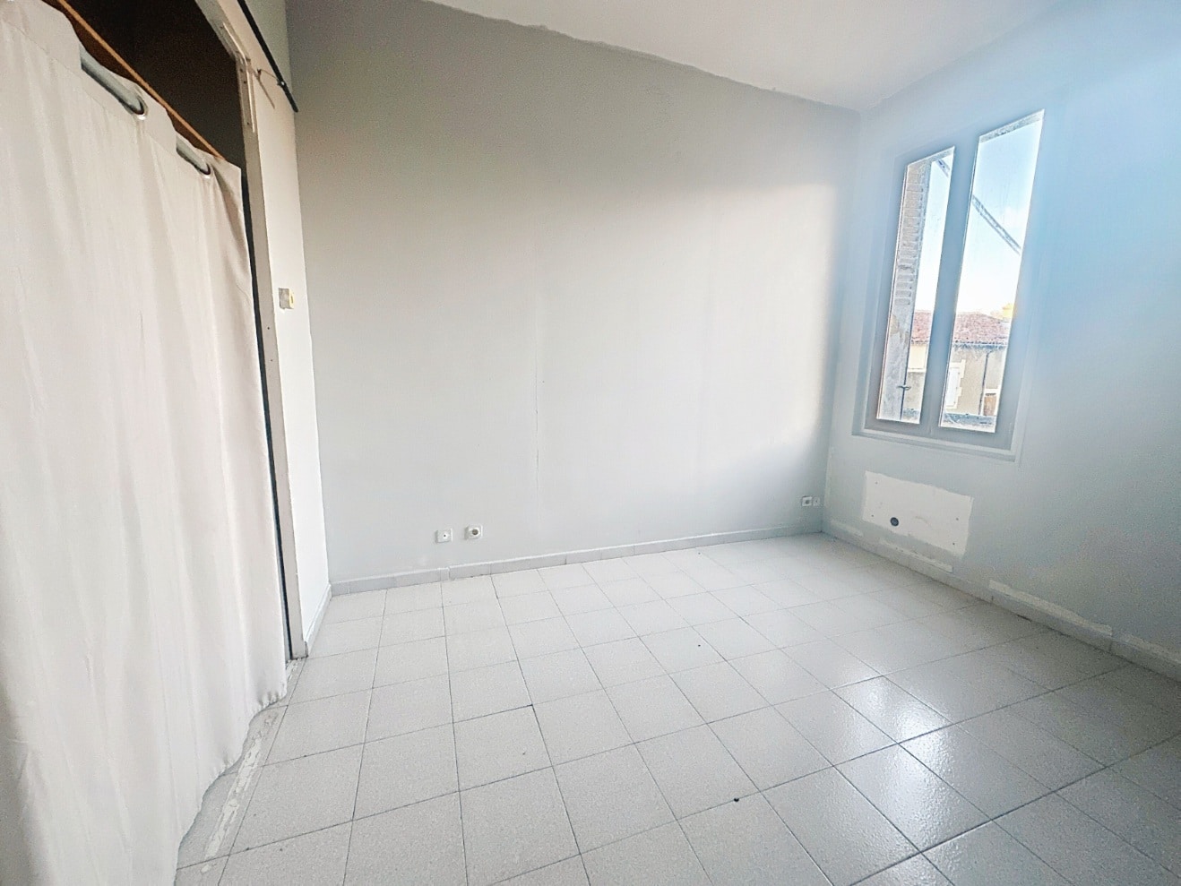Chambre de 12 m² avec carrelage gris au sol et murs blancs. Fenêtre double vitrage en PVC blanc avec vue dégagée et convecteur électrique installé.