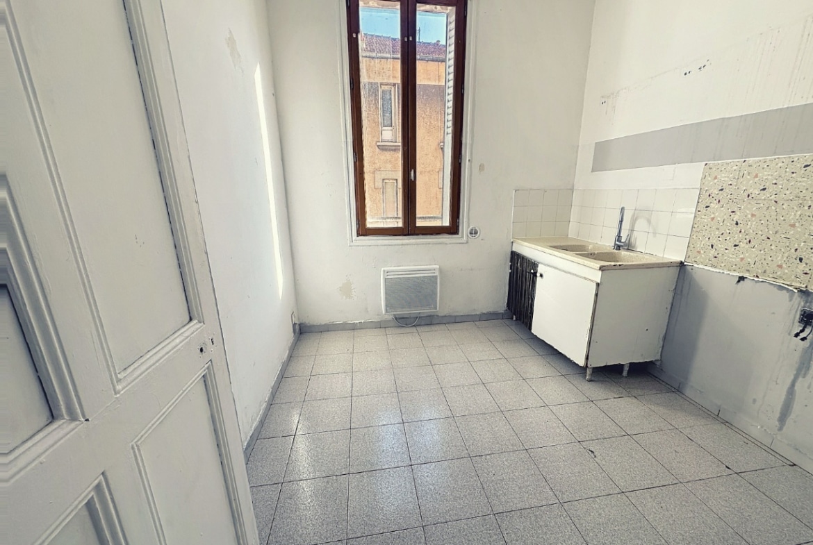 Cuisine de 8 m² avec meuble évier blanc installé et carrelage gris au sol. Fenêtre en bois double vitrage avec volet métallique et convecteur électrique mural