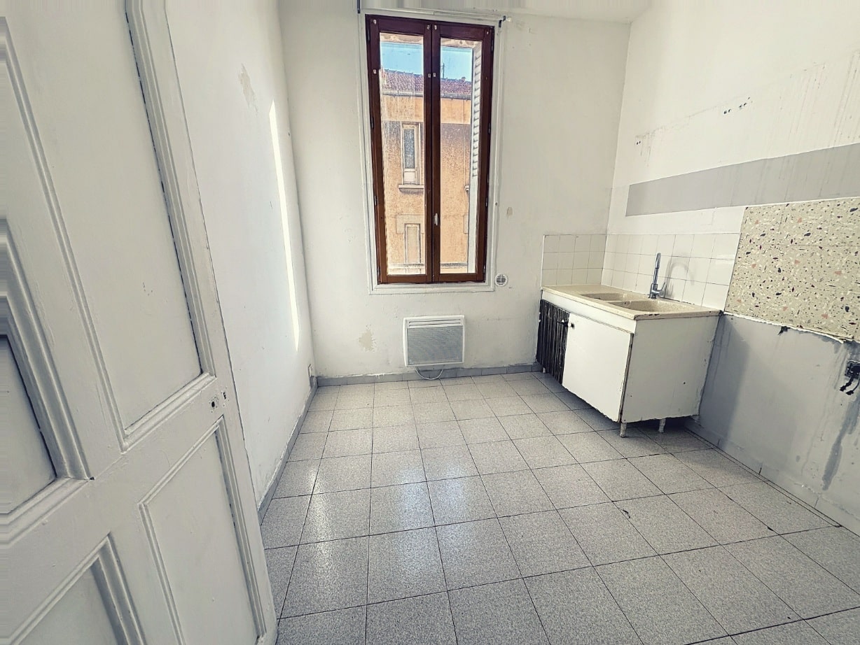 Cuisine de 8 m² avec meuble évier blanc installé et carrelage gris au sol. Fenêtre en bois double vitrage avec volet métallique et convecteur électrique mural