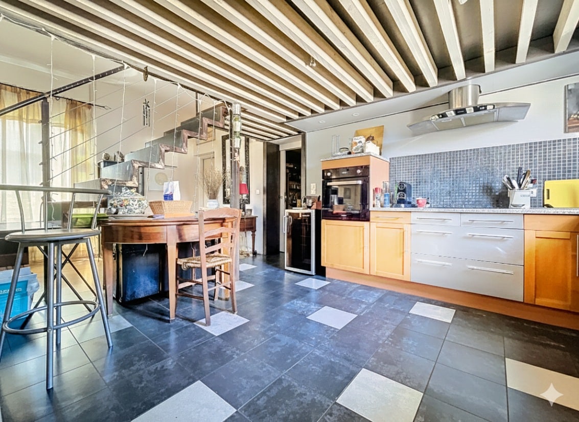 Cuisine aménagée ouverte sur espace repas avec poutres peintes au plafond et carrelage damier au sol. Accès cellier visible avec table ancienne et rangements intégrés dans appartement centre Avignon.