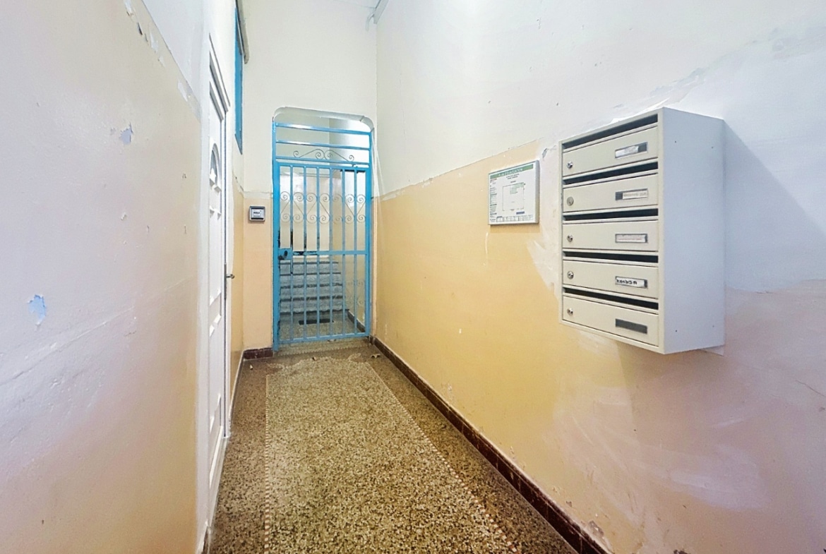 Hall d'entrée de l'immeuble avec interphone et boîtes aux lettres murales. Porte d'accès sécurisée avec grille métallique bleue donnant sur l'escalier intérieur.