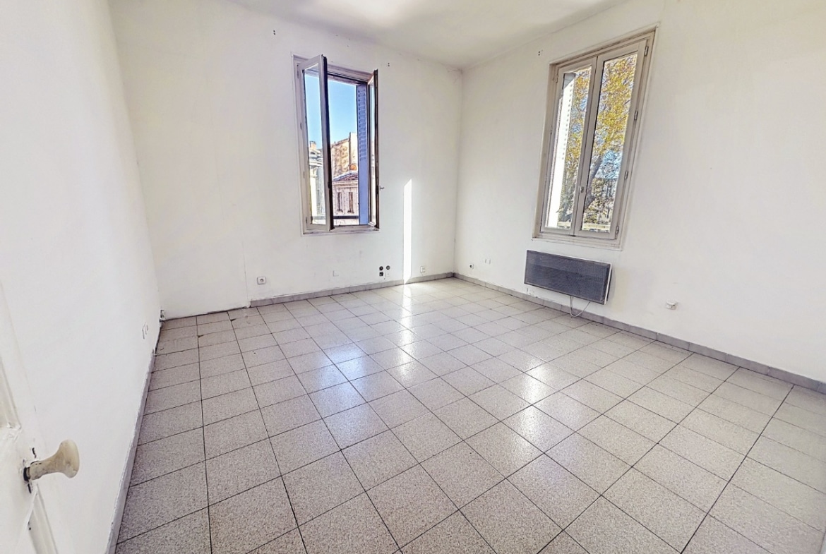 Séjour de 17 m² avec carrelage gris et murs blancs rafraîchis. Deux fenêtres offrant une double exposition avec convecteur électrique et prises électriques apparentes.