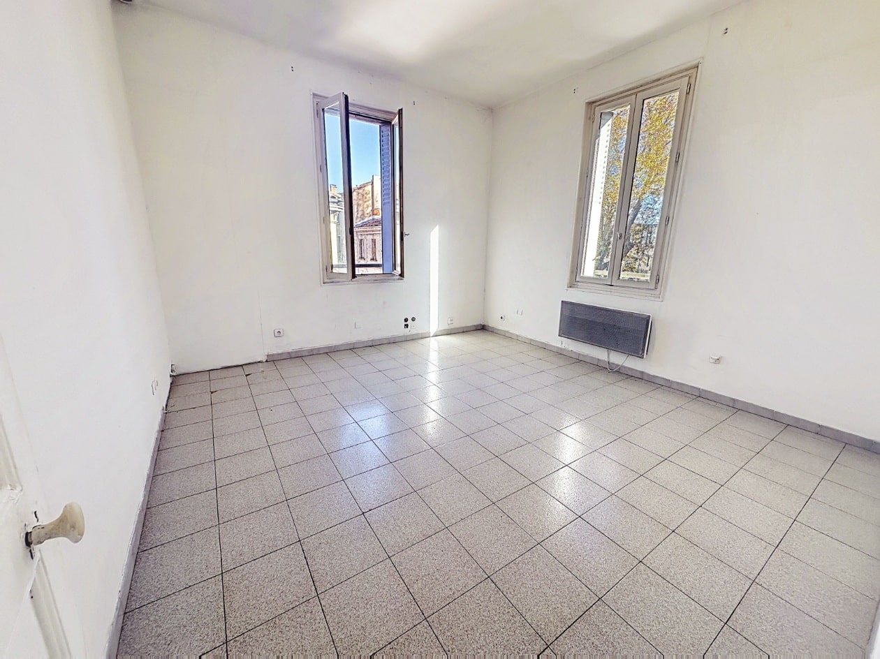 Séjour de 17 m² avec carrelage gris et murs blancs rafraîchis. Deux fenêtres offrant une double exposition avec convecteur électrique et prises électriques apparentes.