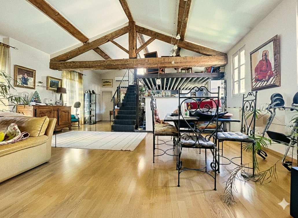 Séjour spacieux avec mezzanine accessible par escalier, poutres apparentes en bois et parquet au sol. Espace de vie traversant avec salle à manger et mobilier sous hauteur de plafond généreuse dans appartement du centre historique d'Avignon.