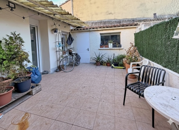 La terrasse est carrelée et comprend plusieurs plantes en pot ainsi qu’un espace pour s’asseoir. Un accès vers la maison et des murs de séparation sont visibles autour de l’espace extérieur.