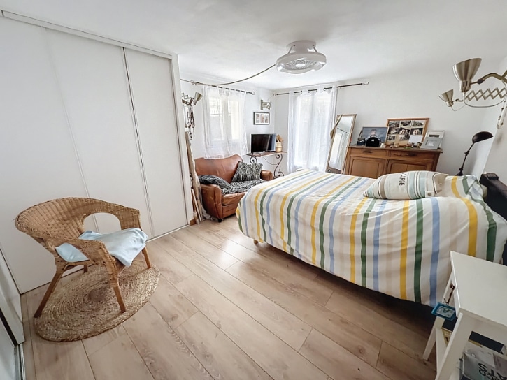 Grande chambre avec lit double, parquet stratifié clair et grands placards coulissants blancs. La pièce bénéficie de deux fenêtres avec rideaux blancs apportant une bonne luminosité naturelle.