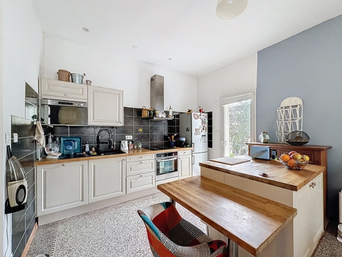 Cuisine en L avec meubles blancs, plan de travail bois, crédence en carrelage noir et hotte inox. Un îlot central avec plateau bois fait office de bar et ouvre sur la salle à manger.