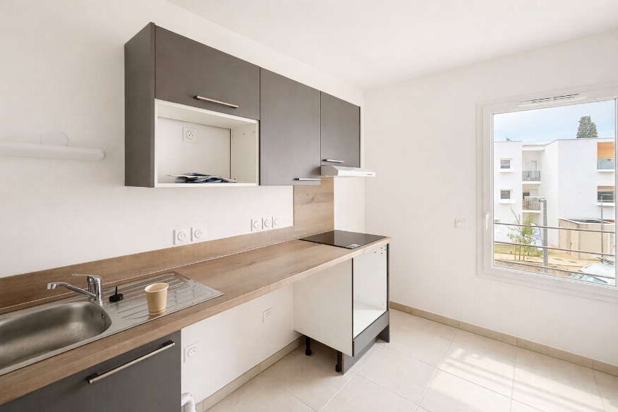 Cuisine ouverte équipée avec meubles gris et plan de travail en bois, plaques de cuisson et hotte intégrées. Appartement T2 neuf en location à Montfavet avec carrelage beige au sol et fenêtres PVC double vitrage donnant sur terrasse