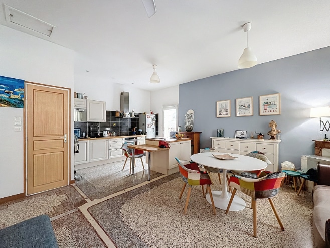 Pièce de vie ouverte avec coin repas, table tulipe et chaises multicolores, cuisine équipée avec îlot central et plan de travail bois. Le mur de fond est peint en bleu gris avec trois cadres décoratifs.