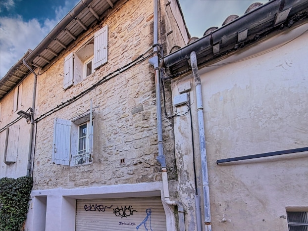 La façade en pierre comporte des fenêtres à volets en bois et des descentes de gouttière visibles. Le bâtiment donne directement sur une rue du centre d’Avignon