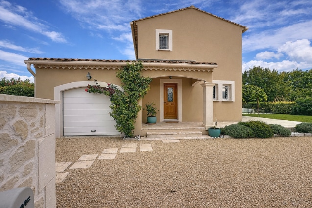 Façade avant de la maison avec garage attenant, porte d'entrée sous porche couvert et terrasse en gravier stabilisé. Enduit beige, volets blancs et toiture en tuiles canal, végétation avec rosiers grimpants sur la façade.