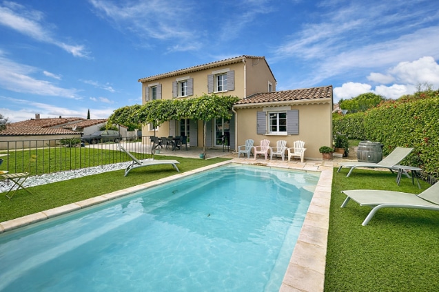 Vue sur la piscine 7x3,5m entourée de pelouse et transats, terrasse couverte avec salon de jardin et chaises longues. Façade arrière avec volets gris, haie de clôture arborée et ciel bleu dégagé.