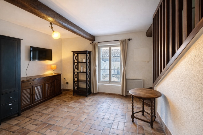 Le salon présente un sol en tomettes, des poutres en bois et une fenêtre apportant de la lumière naturelle. Une armoire, un meuble bas et un escalier sont visibles dans la pièce.