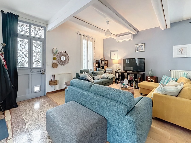 Salon lumineux avec poutres peintes en blanc, canapés bleu et jaune moutarde, parquet clair et climatisation murale. La pièce dispose d'une grande porte vitrée à ferronnerie donnant sur l'extérieur.